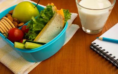 Tupper lleno de frutas y verduras al lado de un vaso de leche y un cuaderno con un lapiz encima